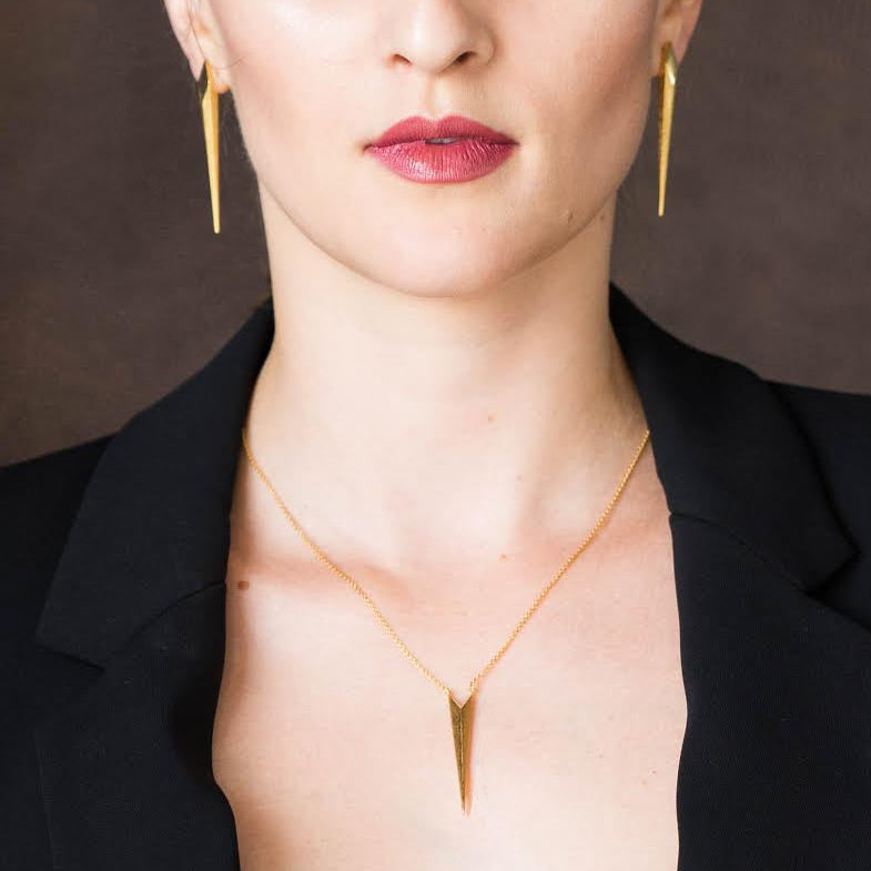 A woman posing in a black blazer with the front open, wearing gold jewelry including a necklace, earrings, and bracelets, set against a dark brown paneled background.