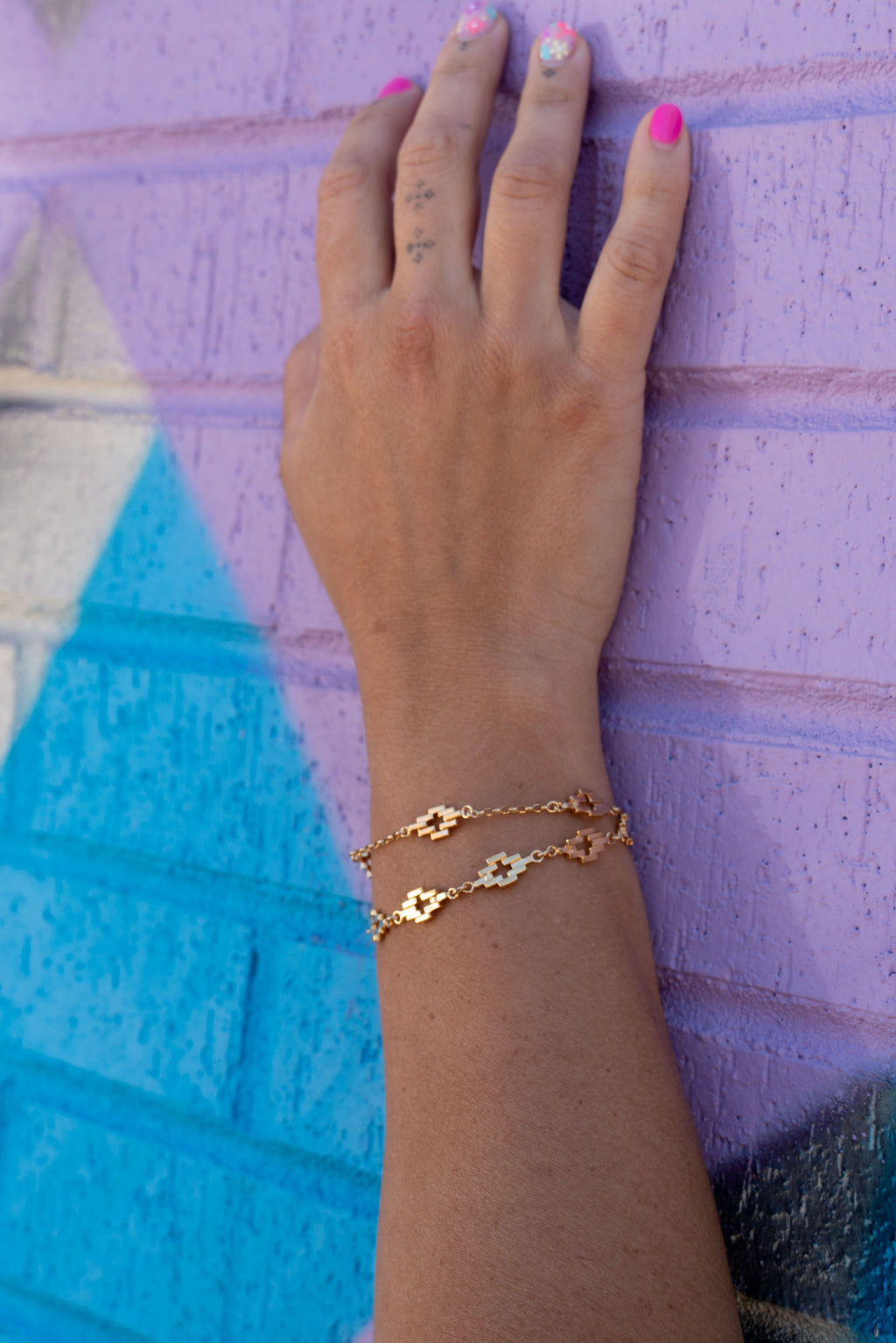Gold Aura Link Bracelet layered with Gold Aura Station bracelet in a wrist against a purple and blue brick background