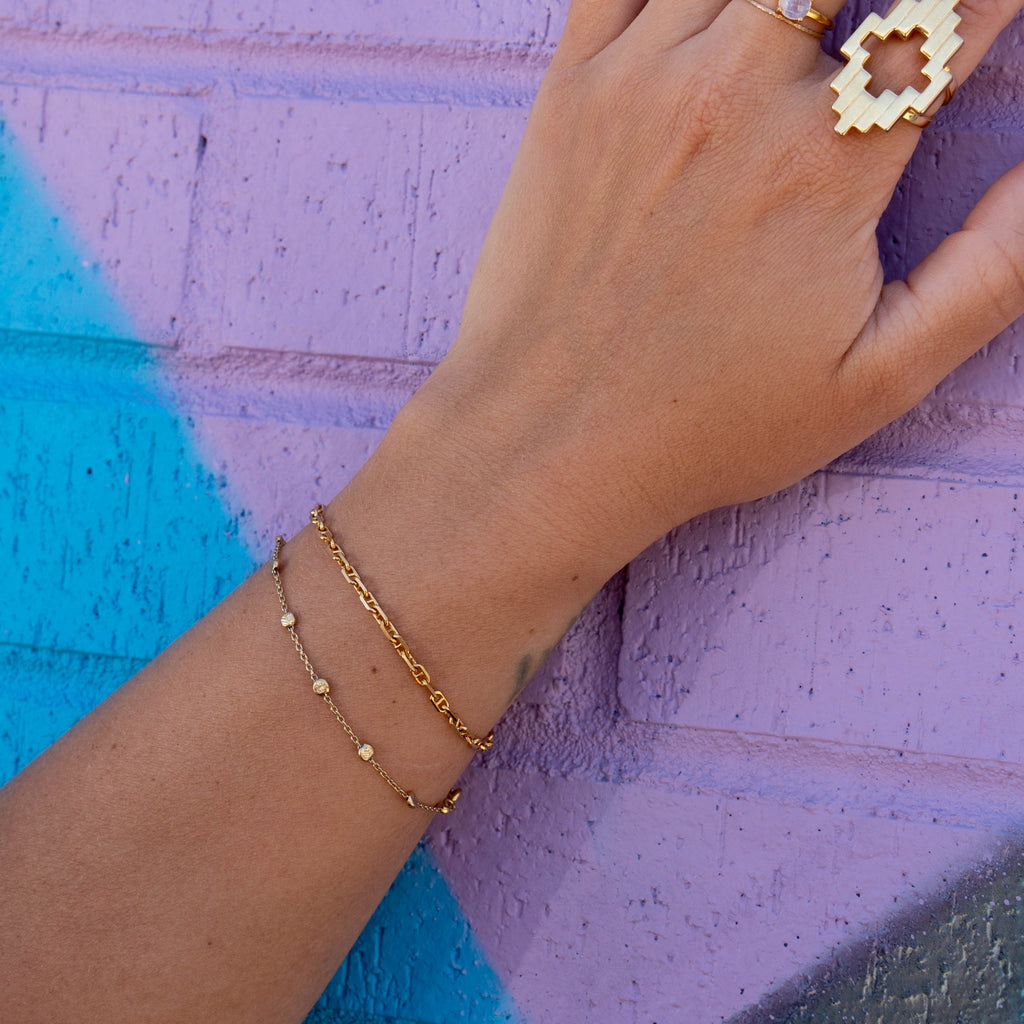 Close up of gold anchor bracelet on wrist layered with the nebula bracelet, also wearing the Aura Ring against purple and blue background