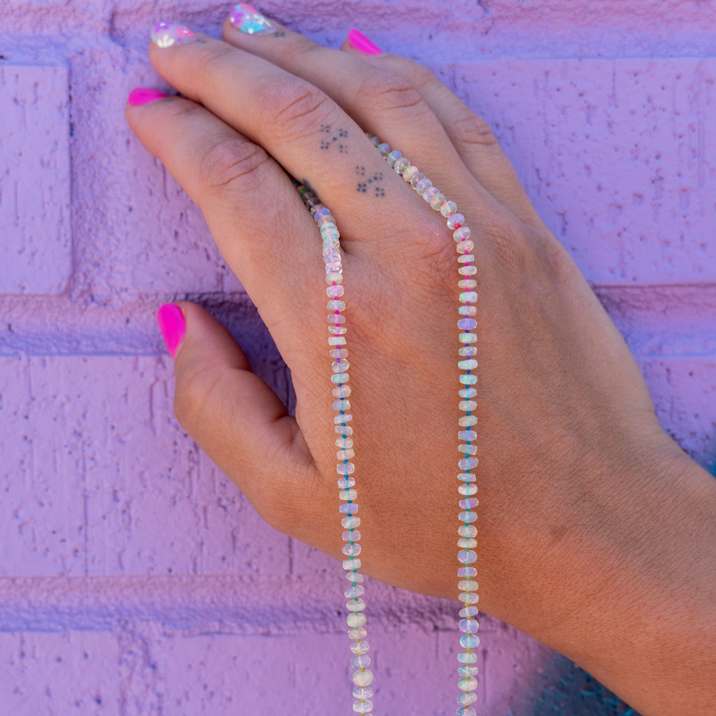 Manicured hand holding 14K Gold and Opal One Of A Kind Necklace on multicolor silk thread, against a purple brick wall