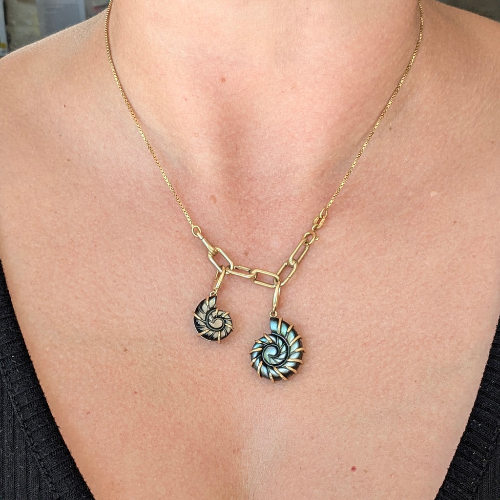A gold chain necklace with a split pendant design, featuring one small spiral shell charm and one larger nautilus-style spiral charm with gold claw setting, worn on a model's neck.