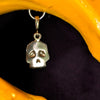 A small gold skull pendant charm hanging from a metal ring, shown against a dark background with a bright yellow curved prop.