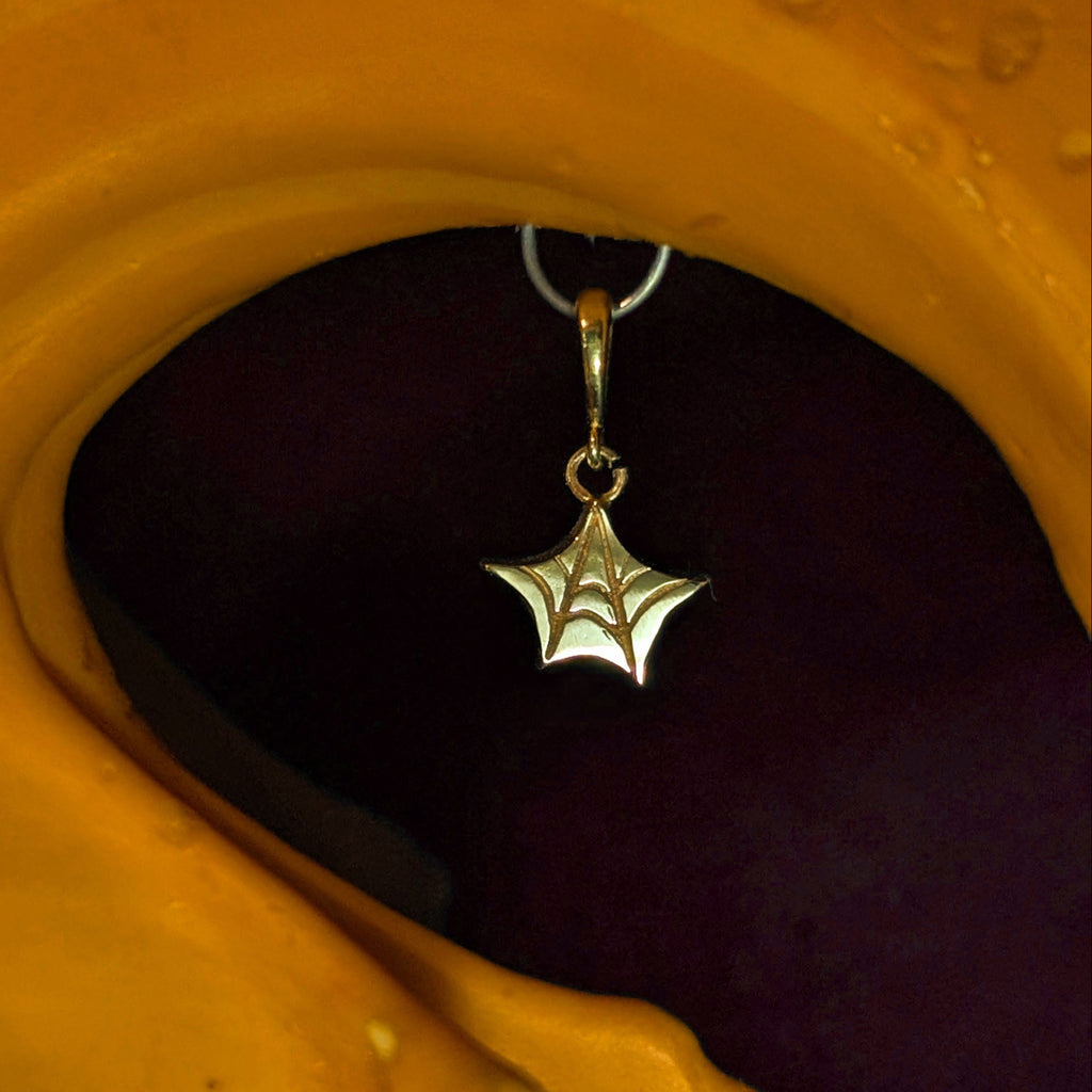A gold spiderweb star-shaped dangle earring hanging from a silver hoop, set against a dark background with a large yellow-orange abstract shape in the foreground.