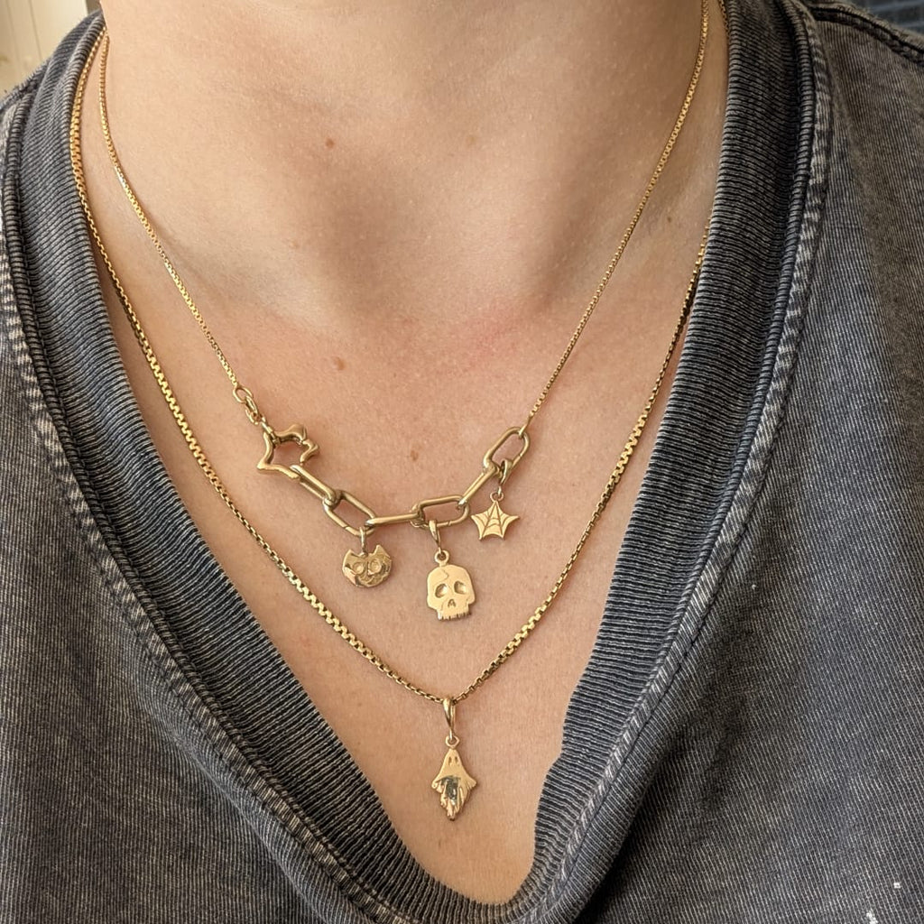 A close-up of a person wearing layered gold necklaces, including a chain necklace with star and cat-shaped charms, a delicate chain necklace with a skull charm, and a longer chain with a small diamond-shaped pendant, styled with a dark gray graphic t-shirt.