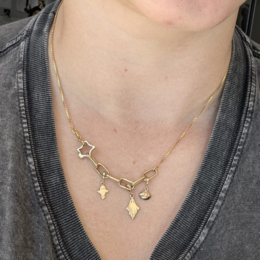 A close-up of a person wearing a delicate gold chain necklace with small dangling charms, including a star, a small heart, and a diamond-shaped pendant, styled over a dark gray V-neck t-shirt.