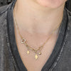 A close-up of a person wearing a delicate gold chain necklace with small dangling charms, including a star, a small heart, and a diamond-shaped pendant, styled over a dark gray V-neck t-shirt.