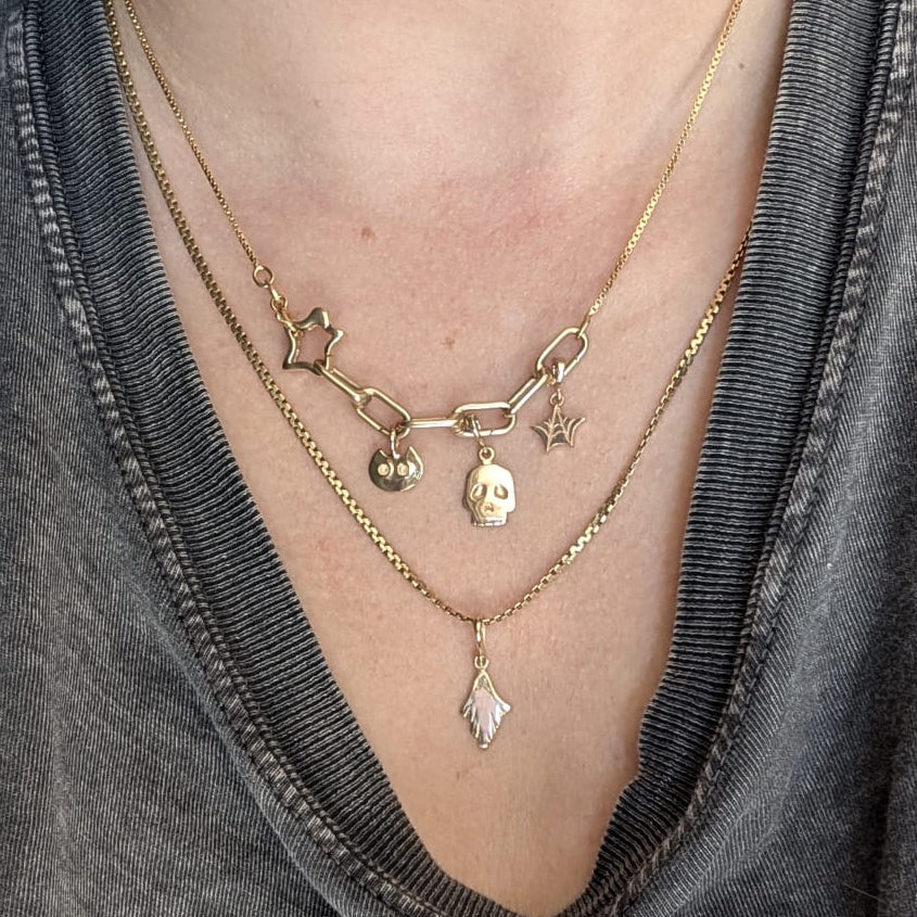 A close-up of a person wearing layered gold necklaces, including a chain necklace with star charms, a link chain necklace with a skull charm, and a delicate chain necklace with a diamond-shaped pendant, styled with a gray graphic t-shirt.