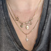 A close-up of a person wearing layered gold necklaces, including a chain necklace with star charms, a link chain necklace with a skull charm, and a delicate chain necklace with a diamond-shaped pendant, styled with a gray graphic t-shirt.