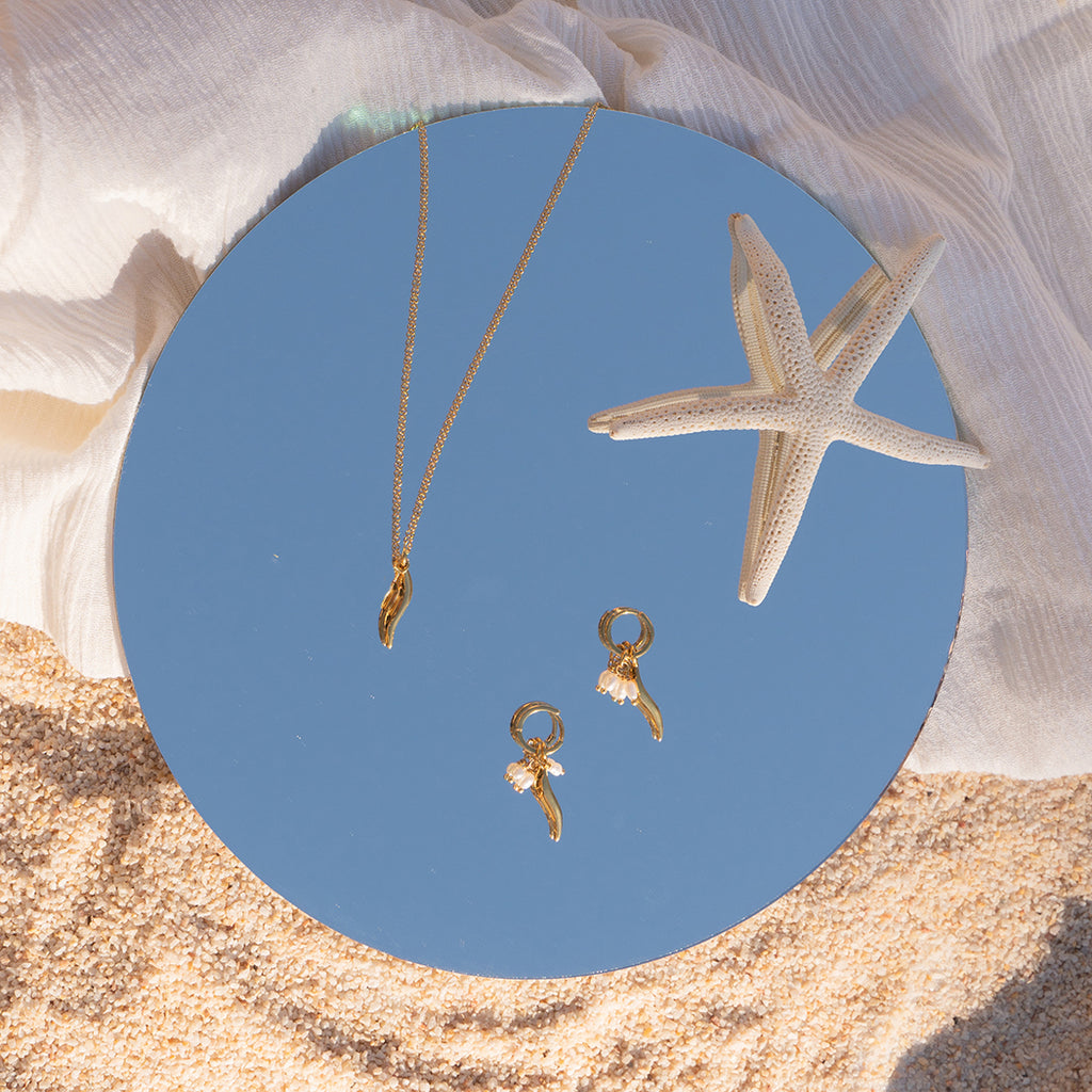 Flat lay of Gold Drift Pearl Huggies, Gold Drift Pendant and starfish on top of mirror with sky reflected in it. The mirror is platedd on top of fabric and sand