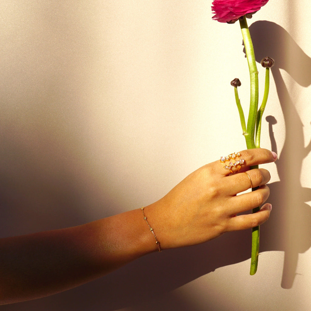 A hand holding a flower and wearing the gold Nebula Bracelet, gold and moonstone via lactea ring and gold Spellbound ring