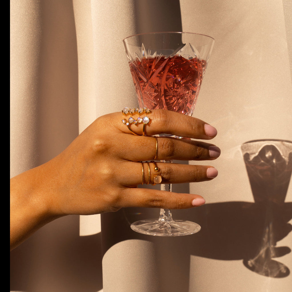 hand holding a glass of wine wearing the Via Lactea Ring in Moonstone and Gold, the Peach moonstone  coeus ring, and gold spellbound rings